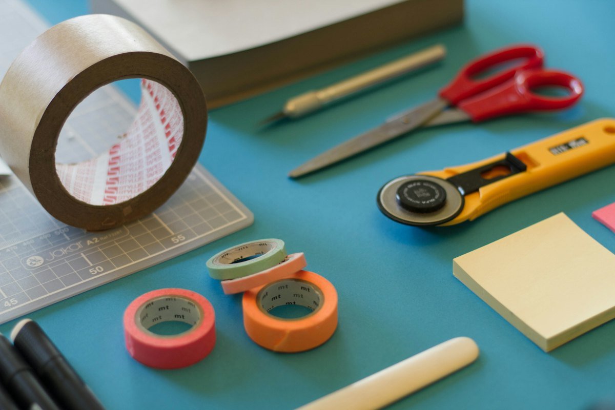 A roll of brown packing tape, scissors, a yellow cutter and small rolls of tape on a blue table